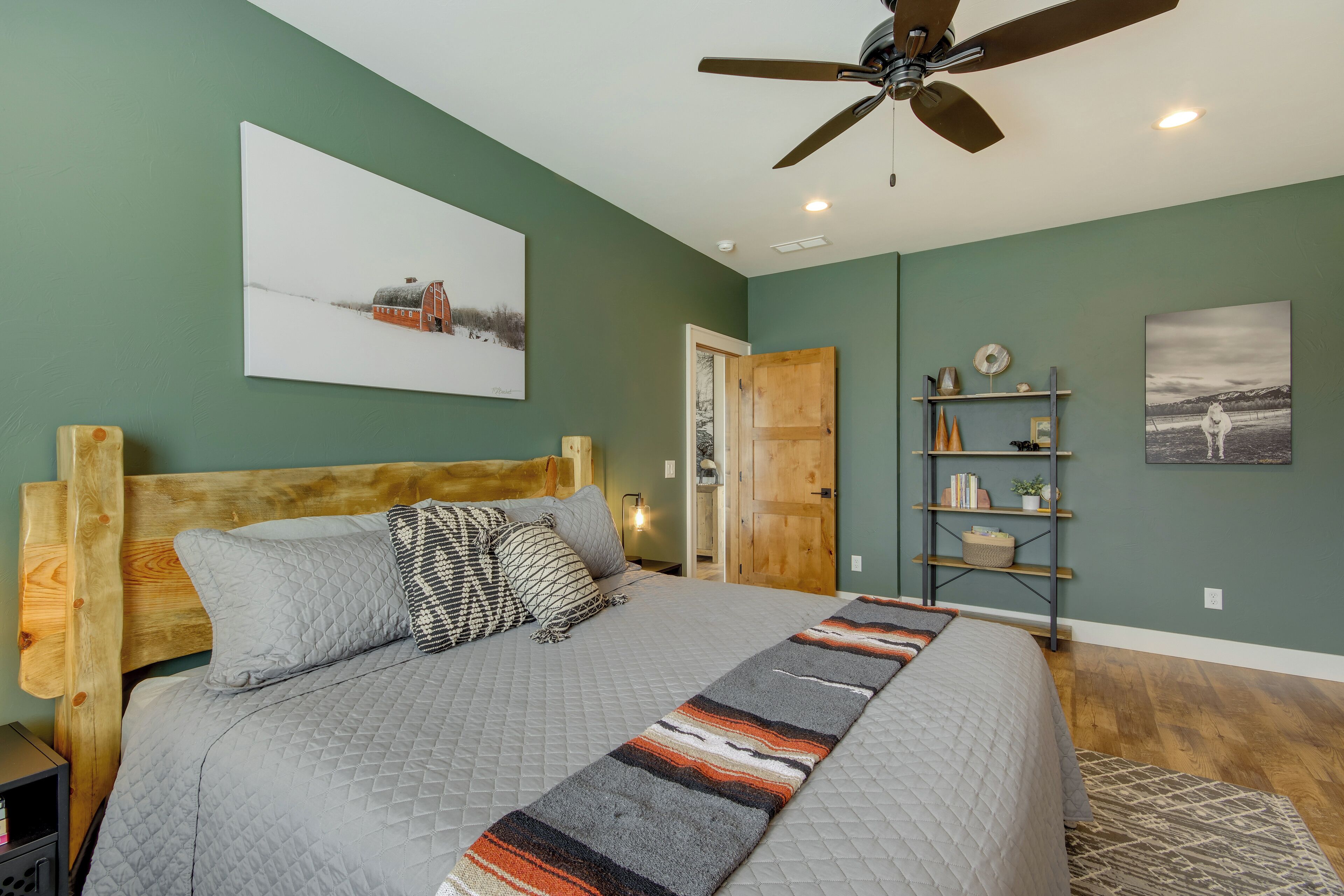 A bedroom with a wooden headboard, a large bed with a quilted gray bedspread, a patterned runner, a ceiling fan, and two wall-mounted shelves with decorative items.