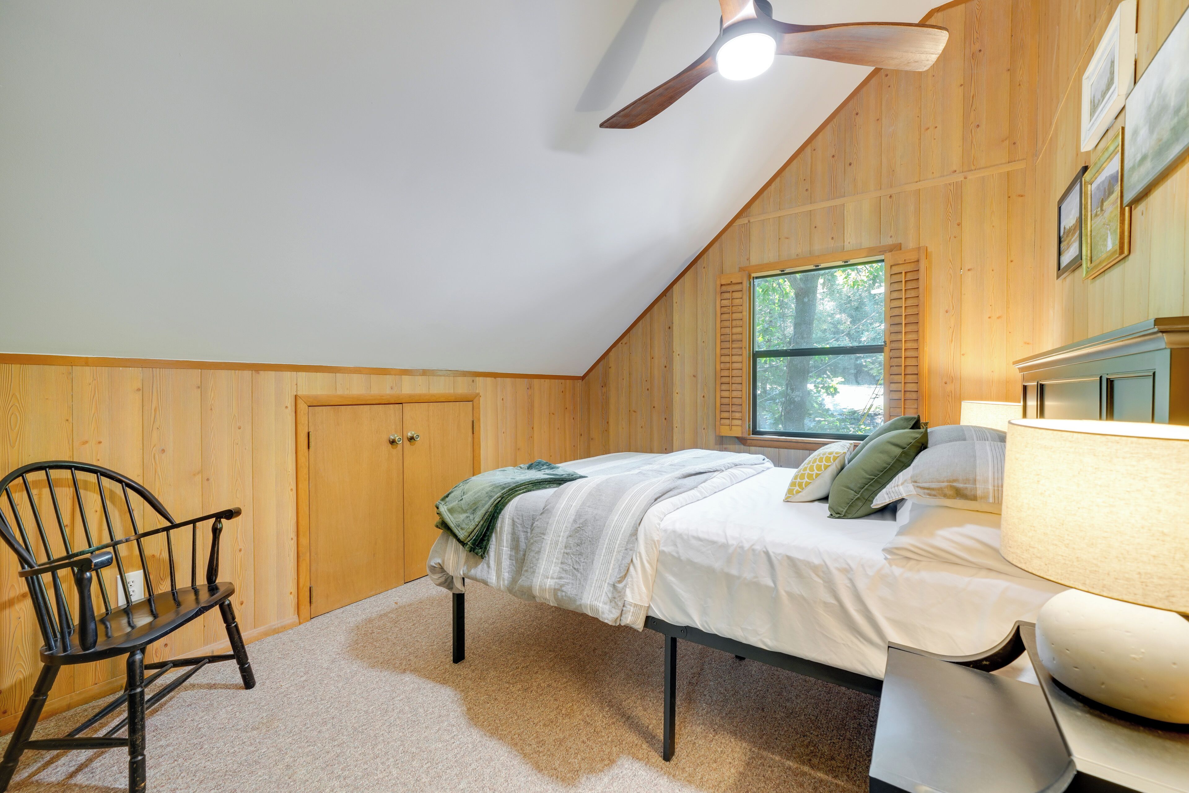 A bedroom with a bed, a chair, a ceiling fan, a window, and wooden walls.