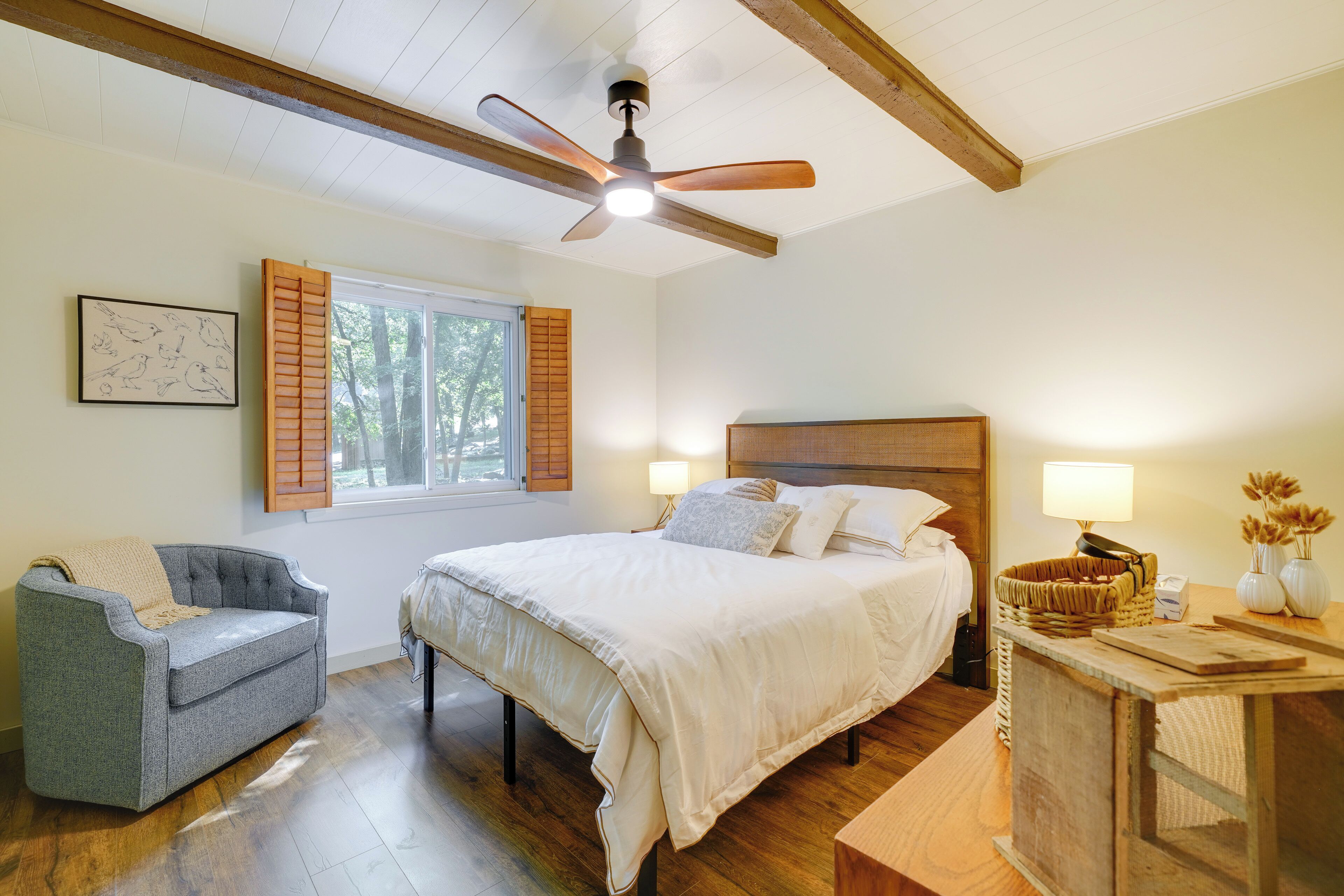 Ein Schlafzimmer mit einem Bett, einem grauen Sessel, einem Holz-Kopfende, einem Deckenventilator und einem Fenster mit Rollläden.