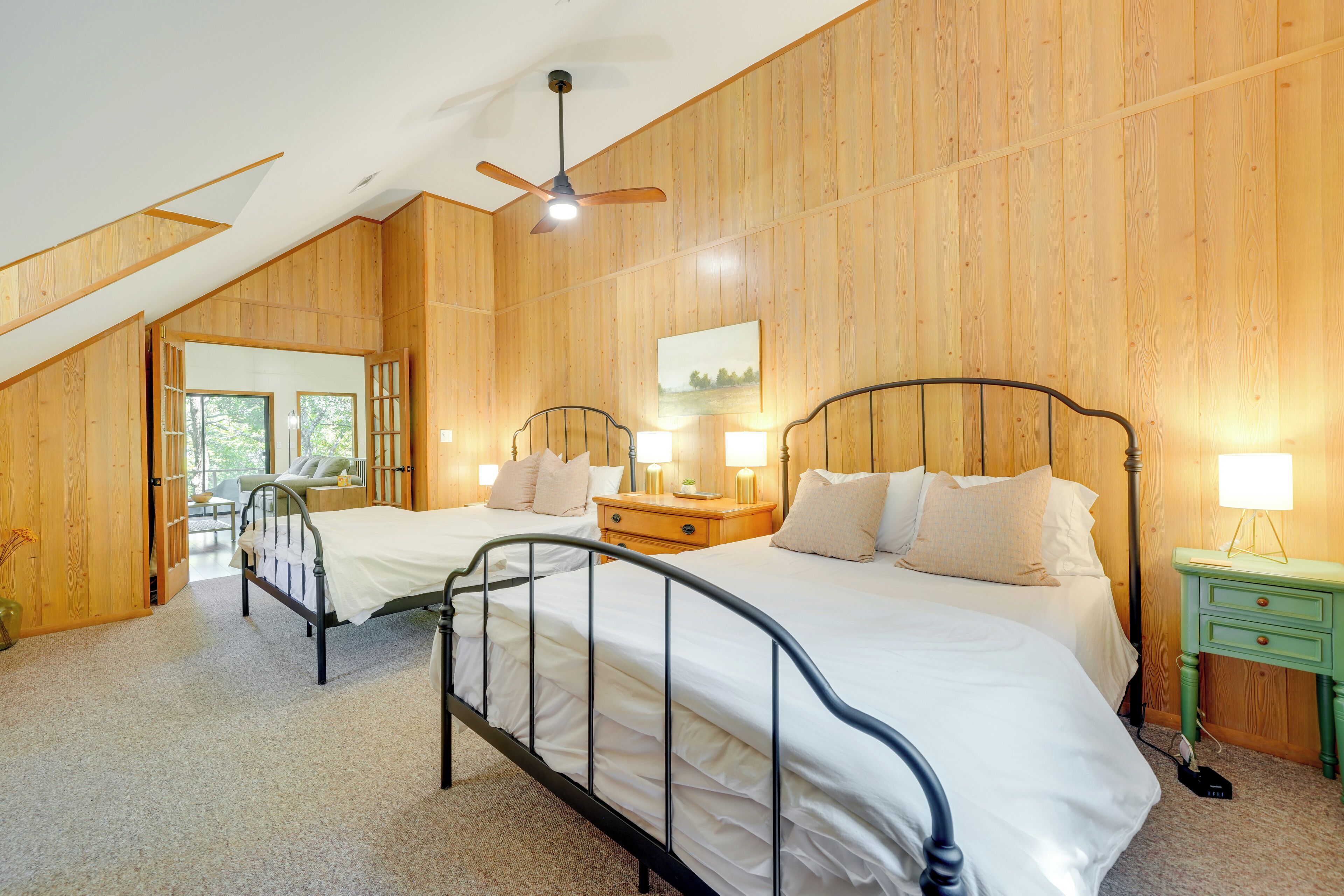 Ein Schlafzimmer mit zwei Betten, Holzwänden, einem Deckenventilator, Nachttischlampen und Blick durch eine offene Tür.