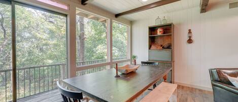 A spacious room with a large wooden table, a bench, and a cushioned chair. There are large windows offering a view of trees.