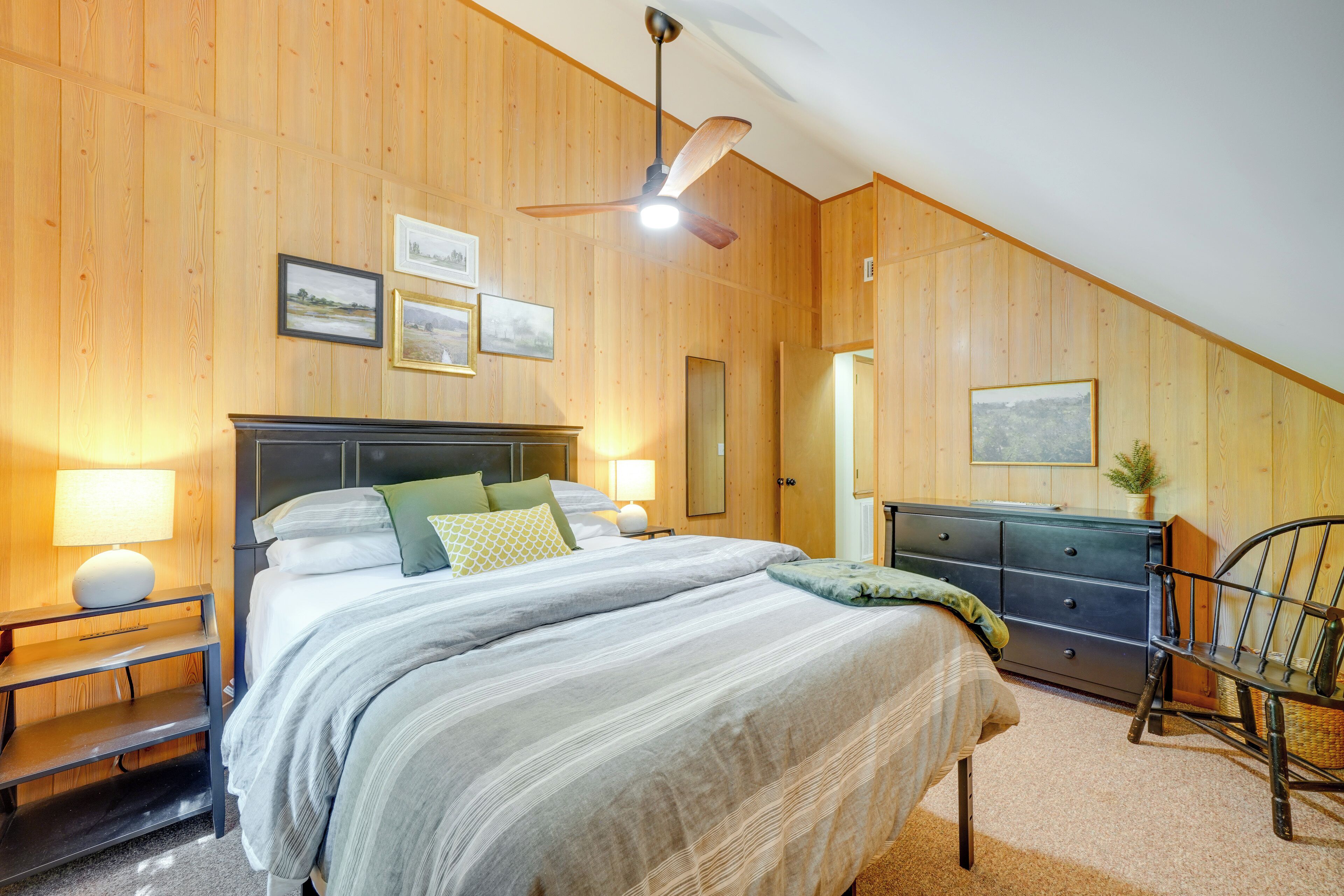 A bedroom with a large bed, wooden walls, a ceiling fan, and a dresser.