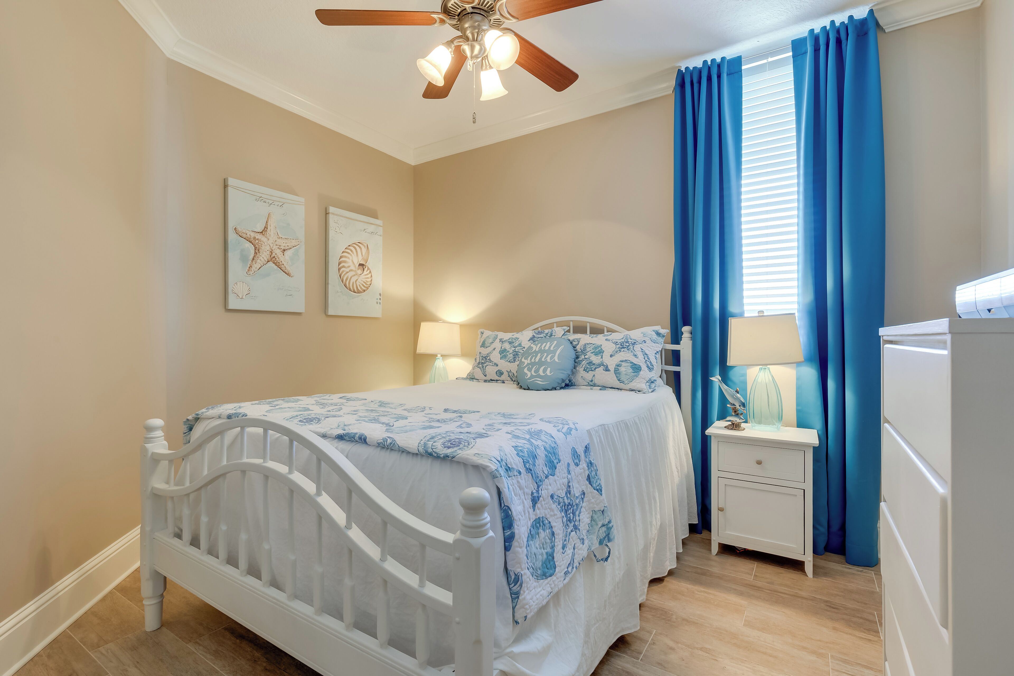 A bedroom with a white bed, blue bedding, a ceiling fan, a nightstand with a lamp, and a window with blue curtains.
