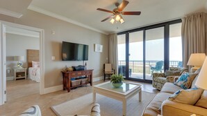 A spacious living room with a ceiling fan, a flat-screen TV, a large window with a view, and a balcony with outdoor furniture. - Dauphin Island Condo in Holiday Isle With Balcony! (Dauphin Island)
