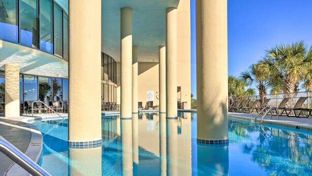 A pool area with clear blue water, surrounded by beige pillars and a modern building with large windows. - Dauphin Island Condo in Holiday Isle With Balcony! (Dauphin Island)