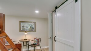 Una habitación con escalera de madera, una mesa redonda con lámpara, un cuadro enmarcado en la pared y una puerta blanca.