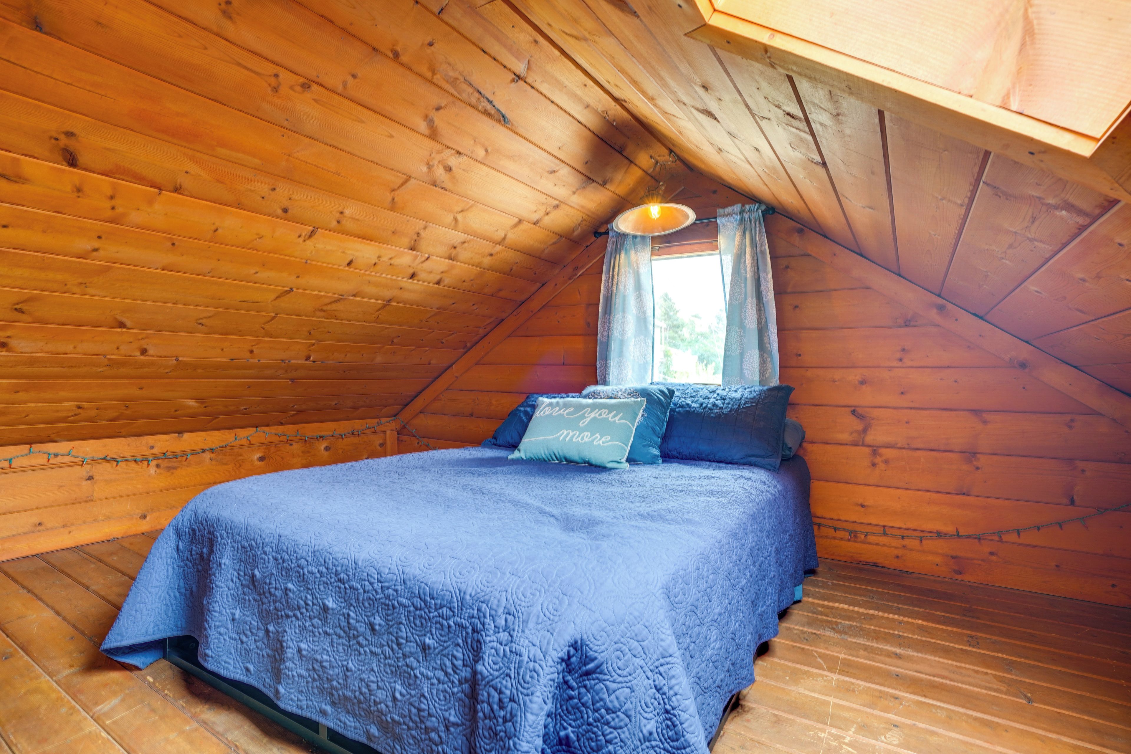 A cozy attic bedroom with a wooden sloped ceiling, a bed with blue bedding, and a window with curtains.