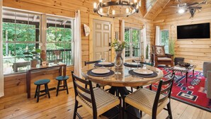 A wooden cabin interior with a dining area, a TV, and a view of the forest. - Pet-friendly Lake Lure Cabin w/ Outdoor Oasis! (Rutherfordton)