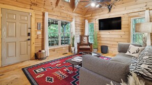 A cozy log cabin interior with a large window, a ceiling fan, a mounted TV, and a comfortable seating area. - Pet-friendly Lake Lure Cabin w/ Outdoor Oasis! (Rutherfordton)