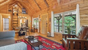 A cozy cabin interior with a dining area, a living room with a sofa and a coffee table, and large windows offering a view of the outdoors. - Pet-friendly Lake Lure Cabin w/ Outdoor Oasis! (Rutherfordton)