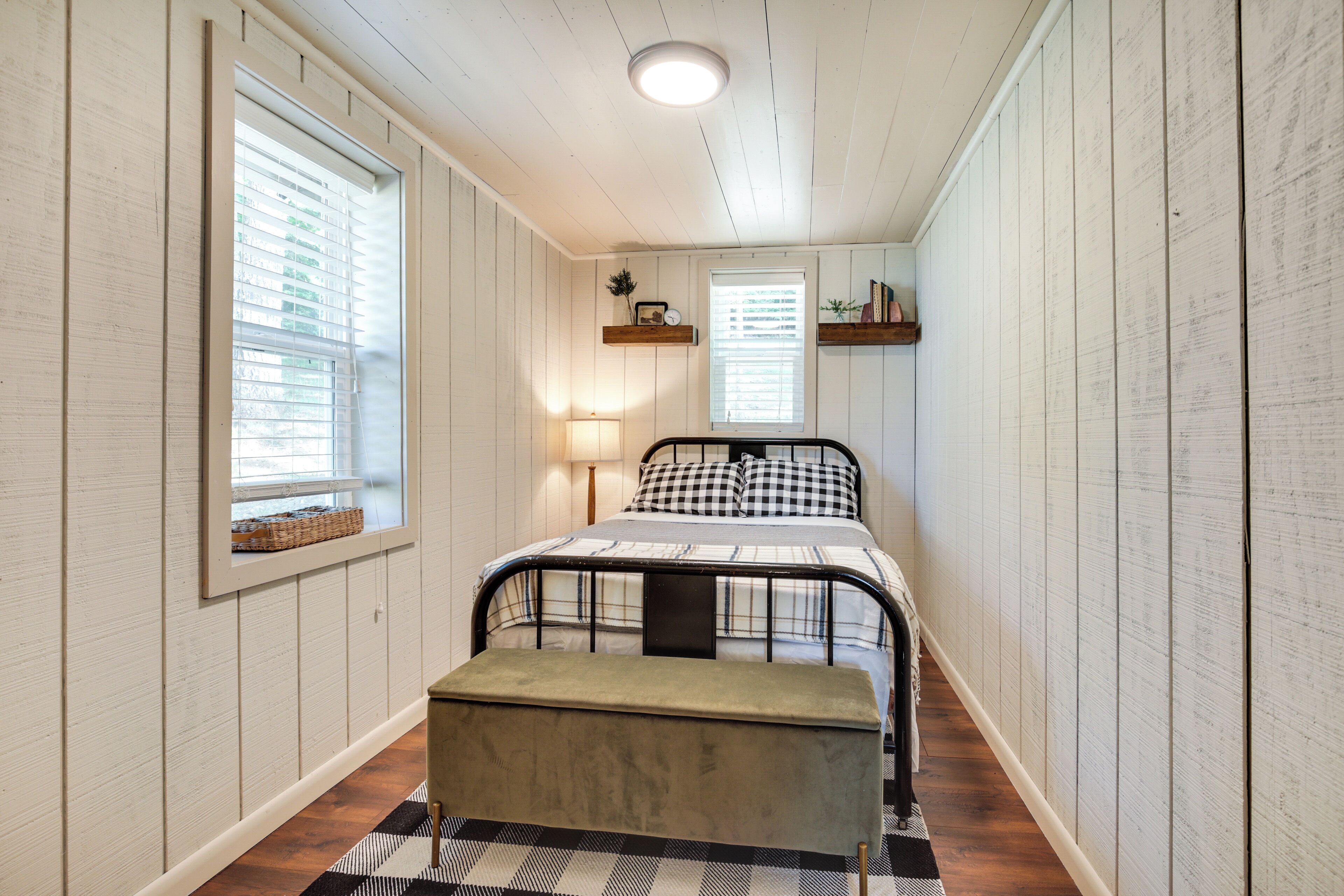 Un dormitorio con cama, mesitas de noche, un banco y dos ventanas con persianas.