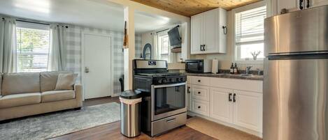 A modern kitchen with a stainless steel refrigerator, a built-in oven, and a microwave. There is a beige sofa and a trash can in the living area.