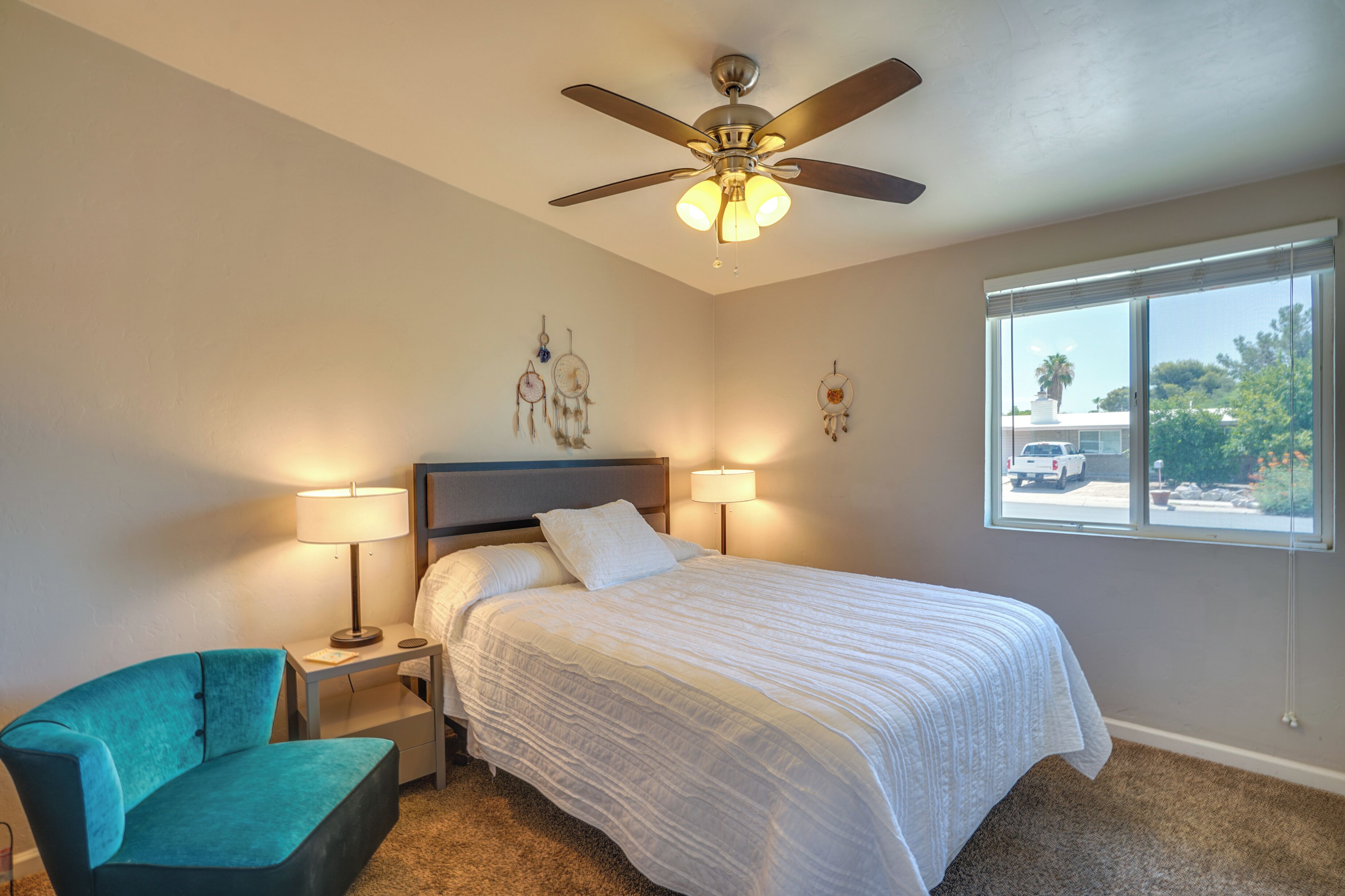 A bedroom with a bed, two bedside lamps, a ceiling fan, a window with a view, and a teal chair.