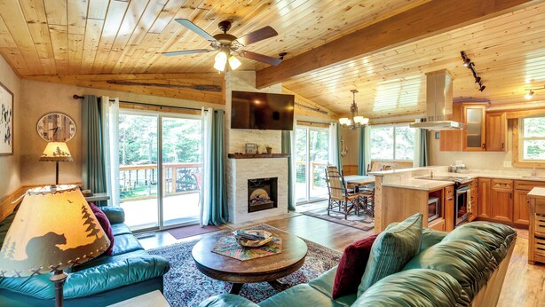 A spacious living room with a fireplace, a ceiling fan, and a large window overlooking a deck. - Waterfront Eagle River Home w/ Dock + Fire Pit! (Eagle River)