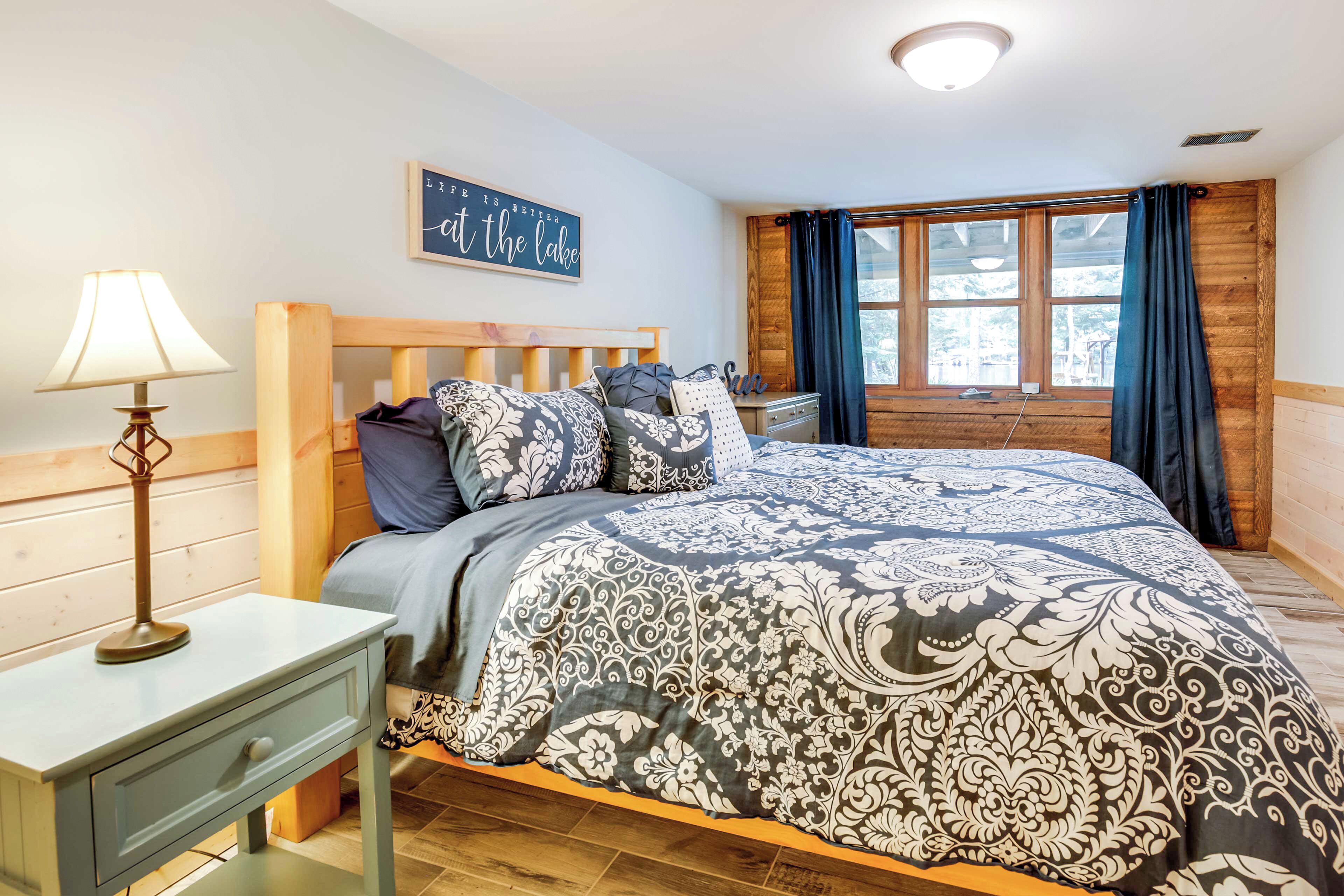 Een slaapkamer met een houten bed, een nachtkastje met een lamp, een raam met gordijnen en een bord aan de muur.