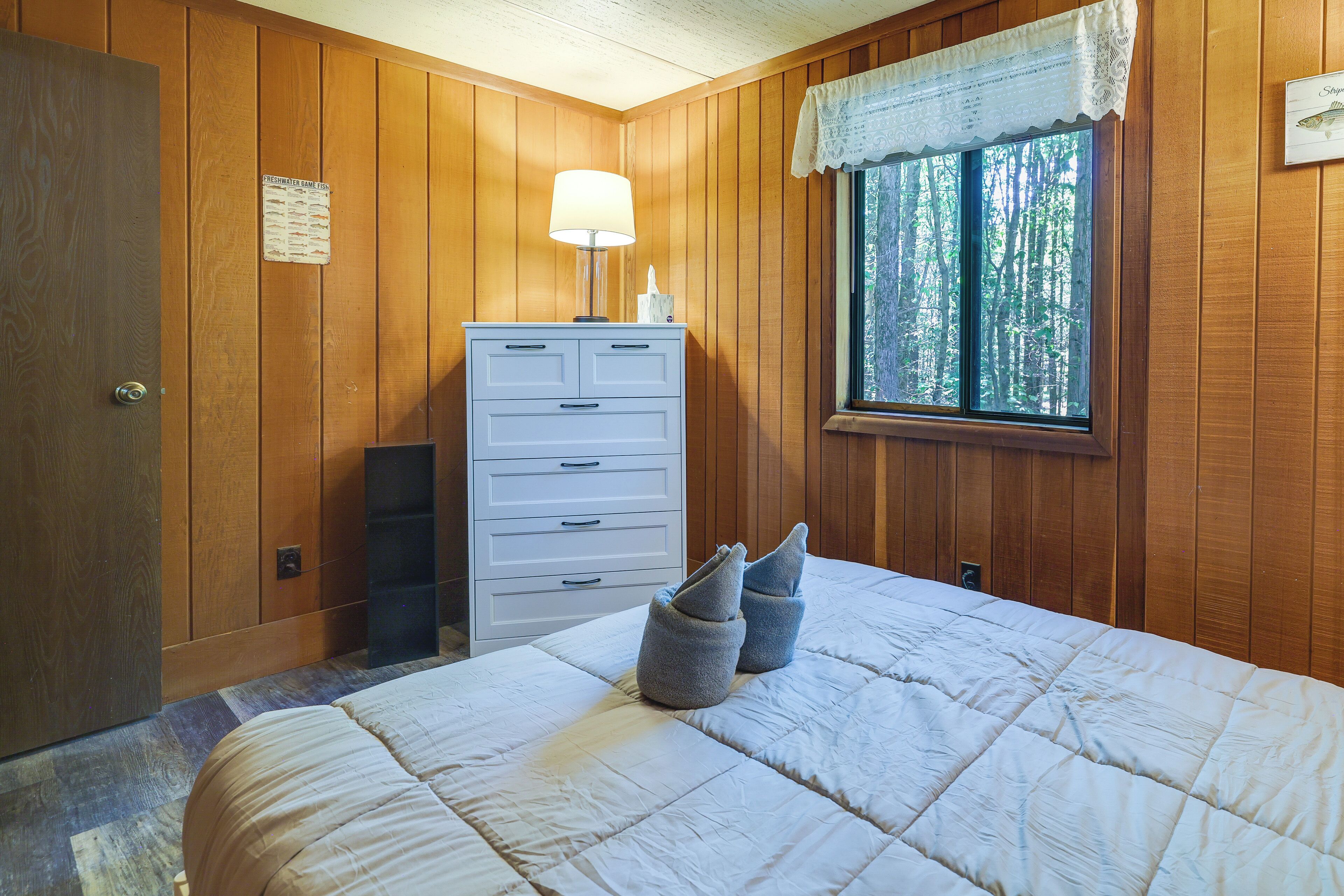 Ein Schlafzimmer mit Holzwänden, einem Bett mit weißer Steudecke, einem Schminktisch und einem Fenster mit Blick auf Bäume. Eine Lampe steht auf dem Schminktisch.