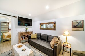 A living room with a brown leather sofa, a wooden side table with a lamp, a bookshelf, and a television mounted on the wall. - Kitty Hawk Vacation Rental w/ Pool (Kitty Hawk)