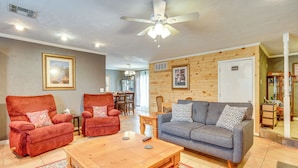 A living room with a wooden ceiling, a ceiling fan, a grey sofa, two red armchairs, a wooden coffee table, and a patterned rug.