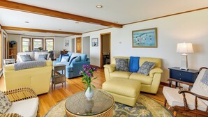 A spacious living room with a yellow sofa, a blue sofa, and a yellow armchair. There is a glass coffee table with a vase of flowers. - Waterfront Massachusetts Vacation Rental w/ Deck (Westport Point)