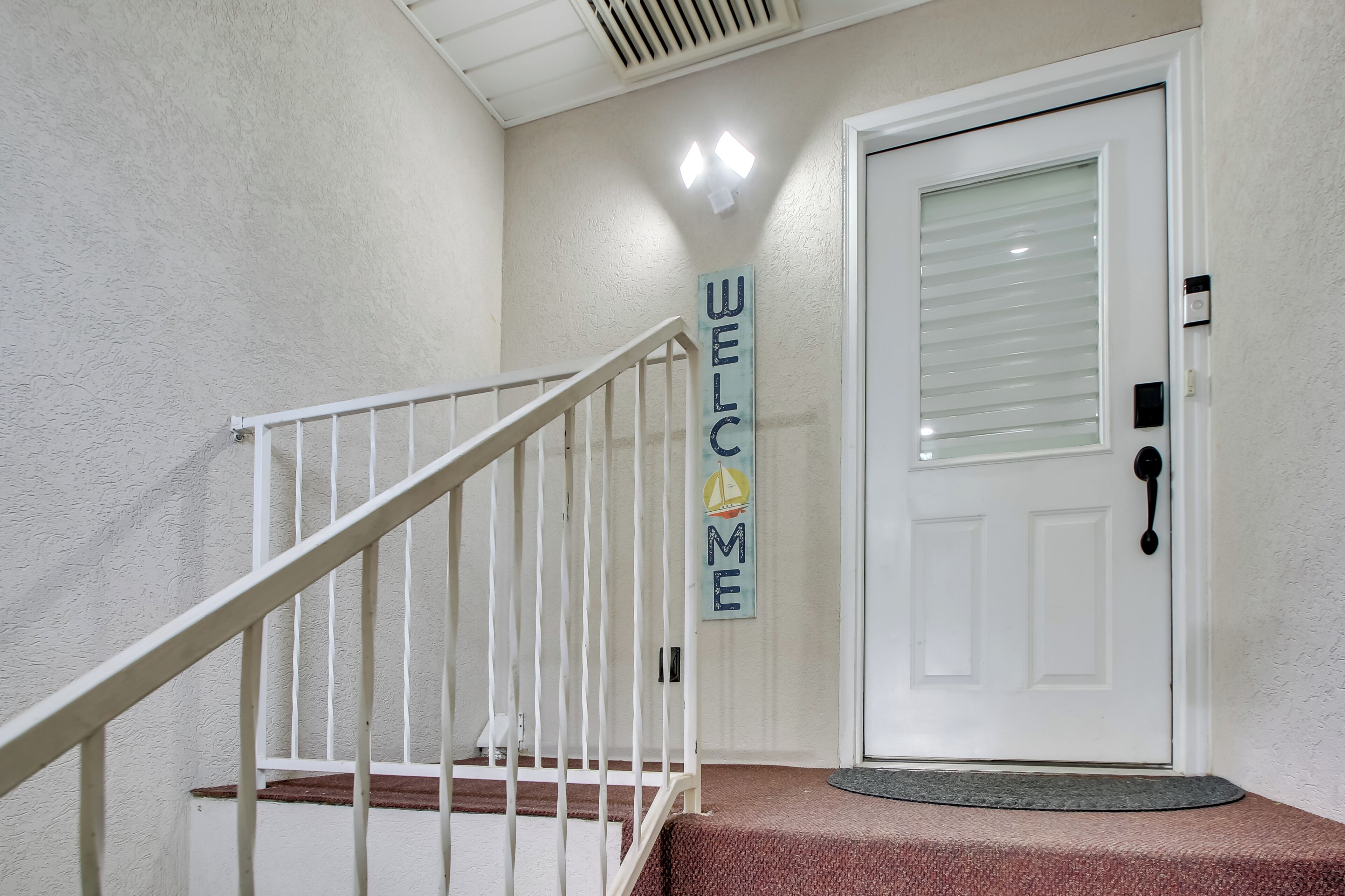 A white door with a 'WELCOME' sign, a white staircase with a red carpet, and a textured wall.