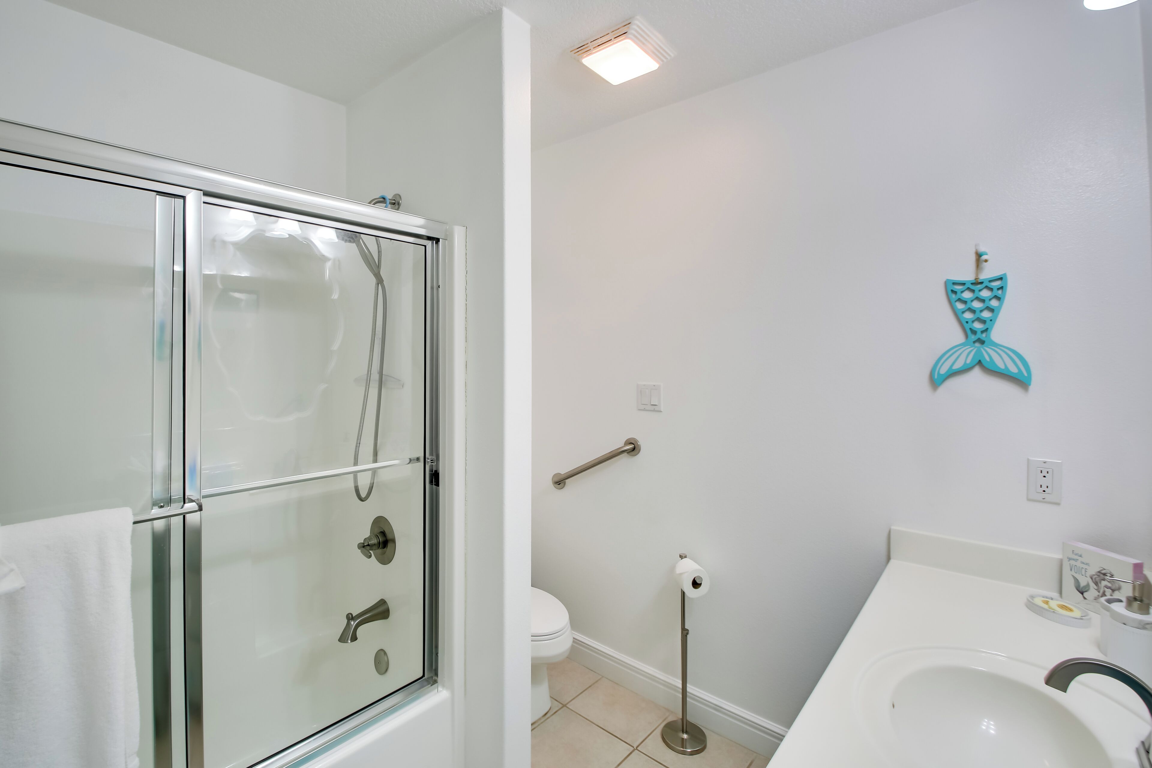 A bathroom with a glass-enclosed shower, a toilet, a sink, and a decorative mermaid tail on the wall.