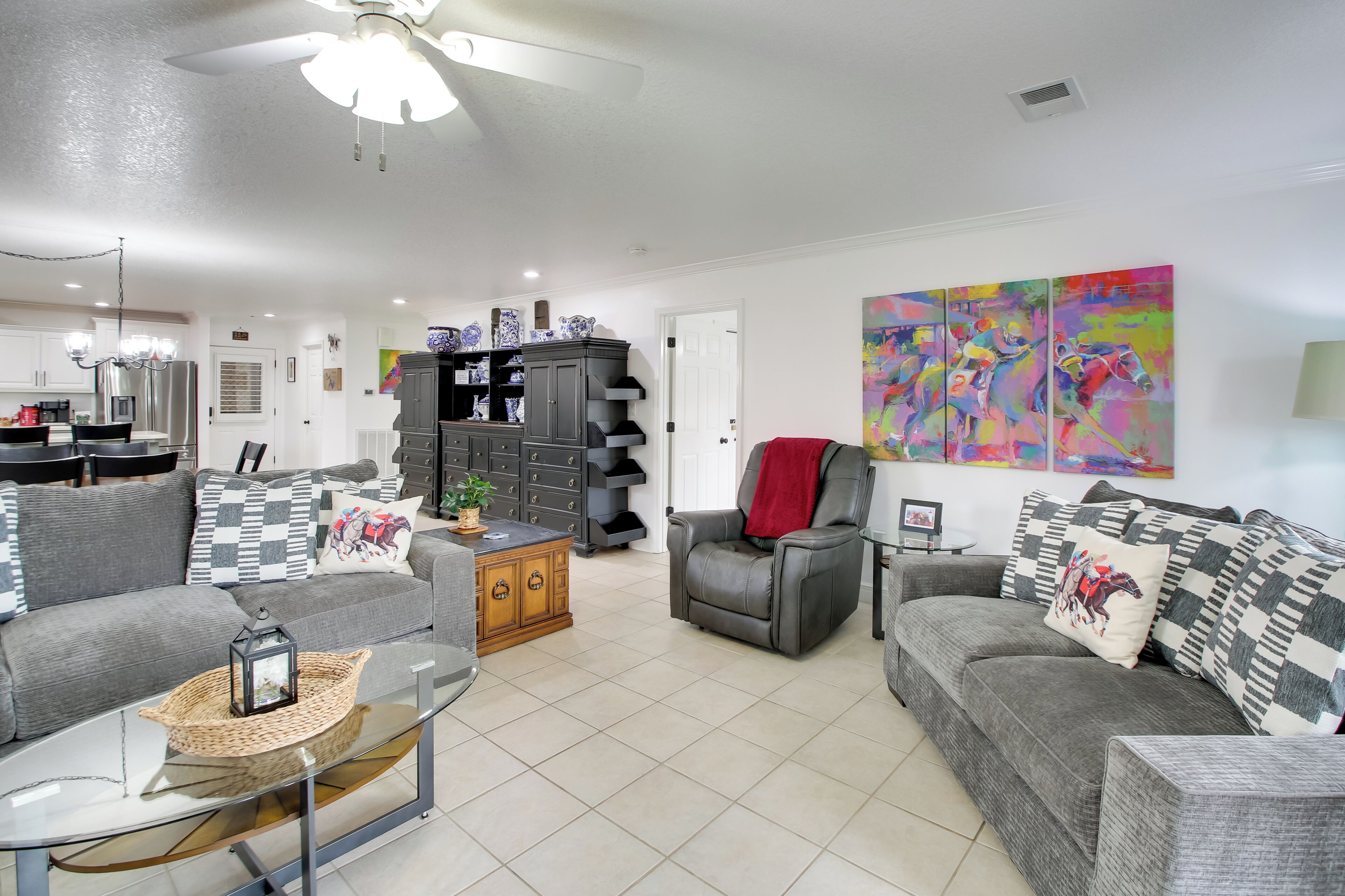 A living room with a sofa, armchair, and a coffee table. There is a kitchen area in the background with cabinets and a refrigerator.