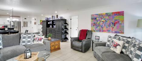 A living room with a sofa, armchair, and a coffee table. There is a kitchen area in the background with cabinets and a refrigerator.