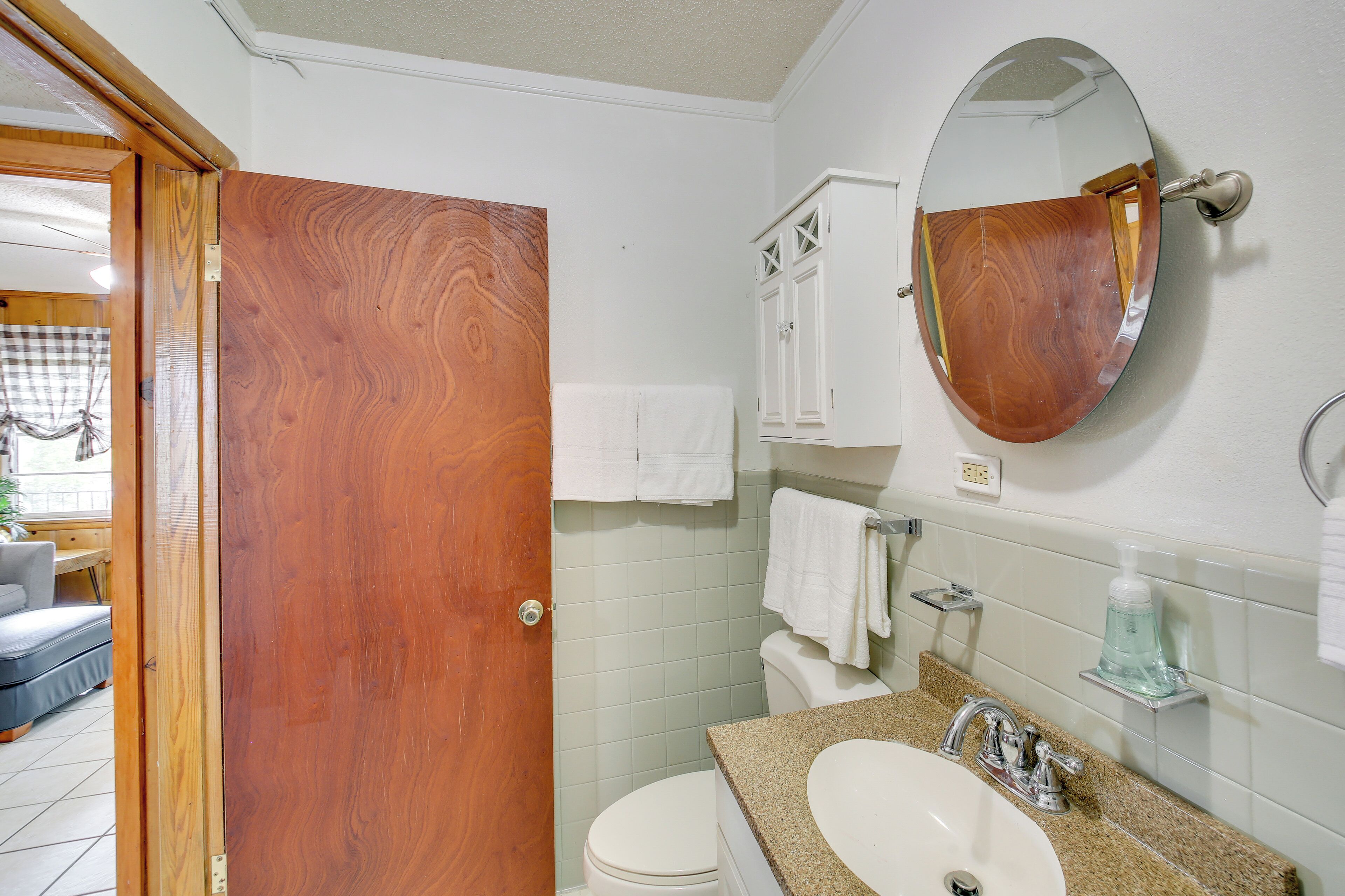Un baño con puerta de madera, inodoro blanco y lavamanos con espejo.
