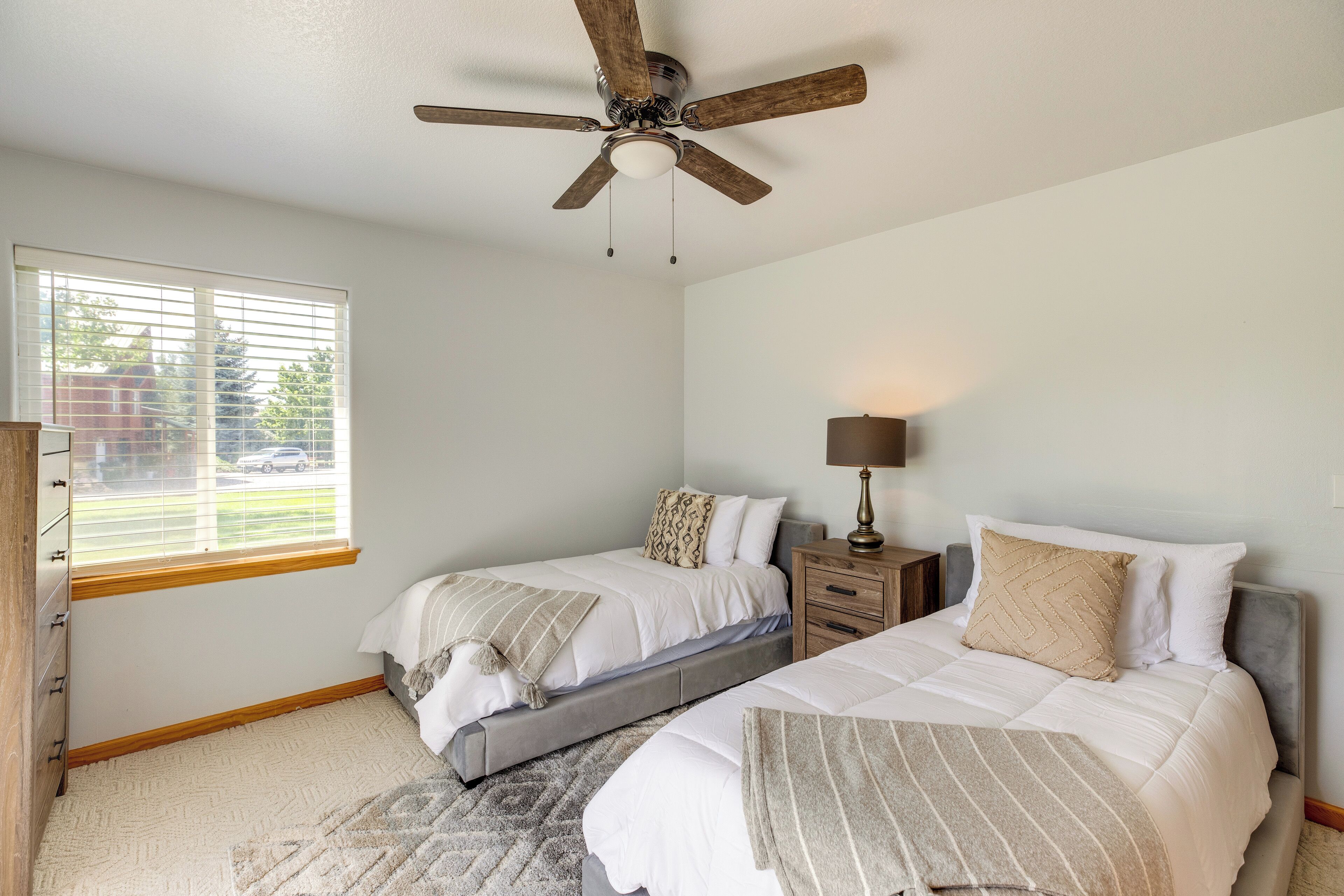 A bedroom with two beds, a ceiling fan, a window with blinds, a nightstand with a lamp, and a dresser.