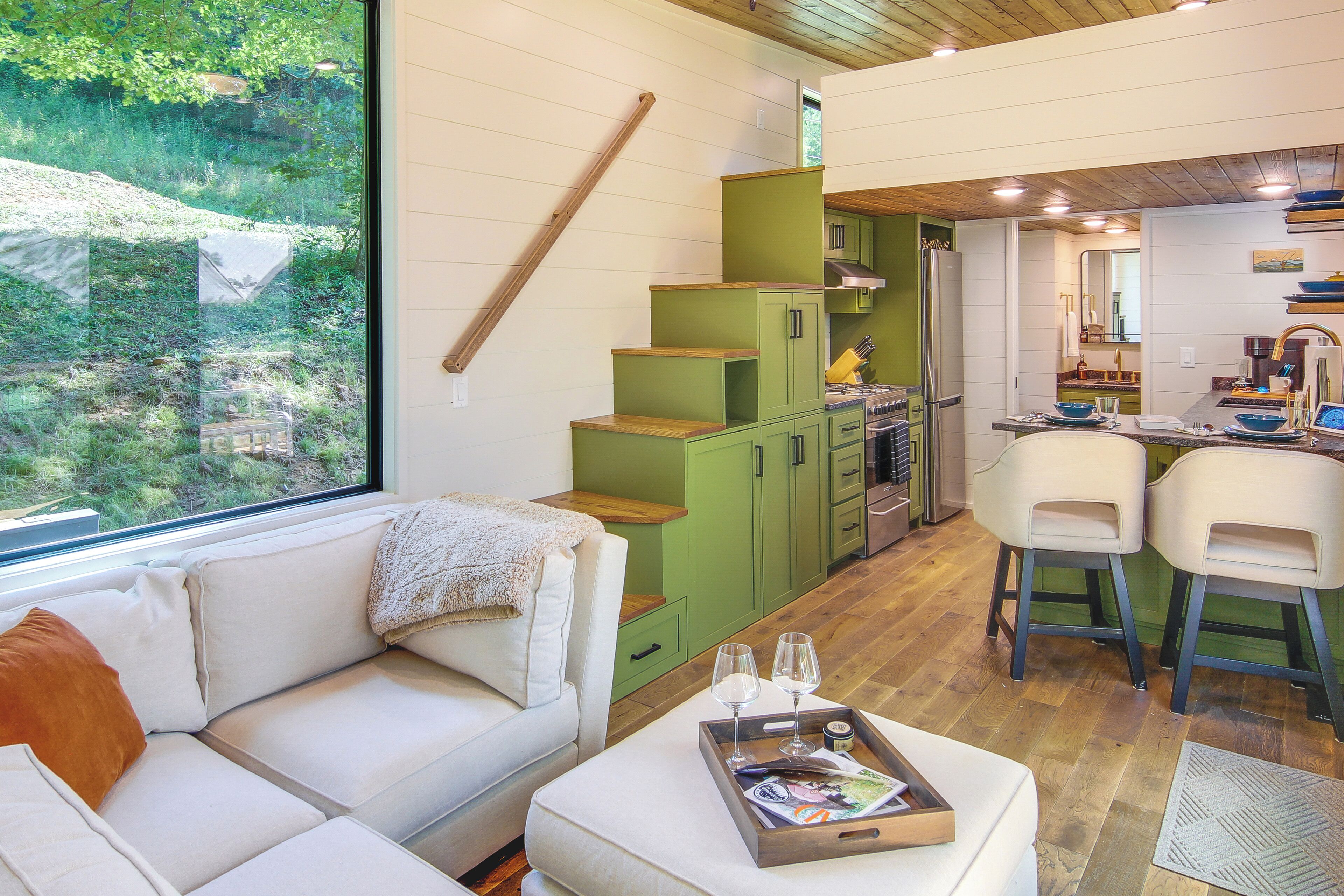 A modern interior with a sofa, a dining area with two chairs, a kitchen with green cabinets, and a large window overlooking greenery.