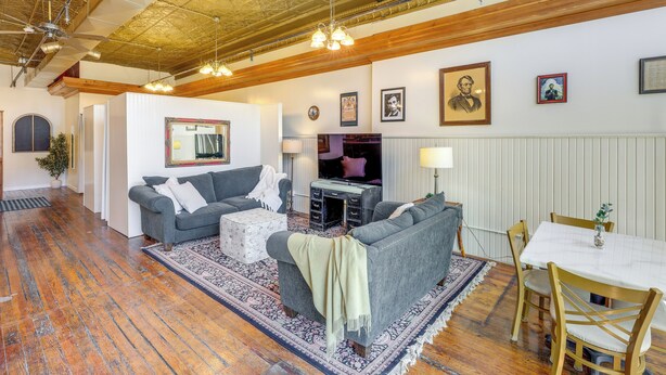 A spacious living room with hardwood floors, a grey sofa, a coffee table, and a dining area with wooden chairs and a white tablecloth. - Cozy Abode in Historic Yankton - Heart of the City (Yankton)