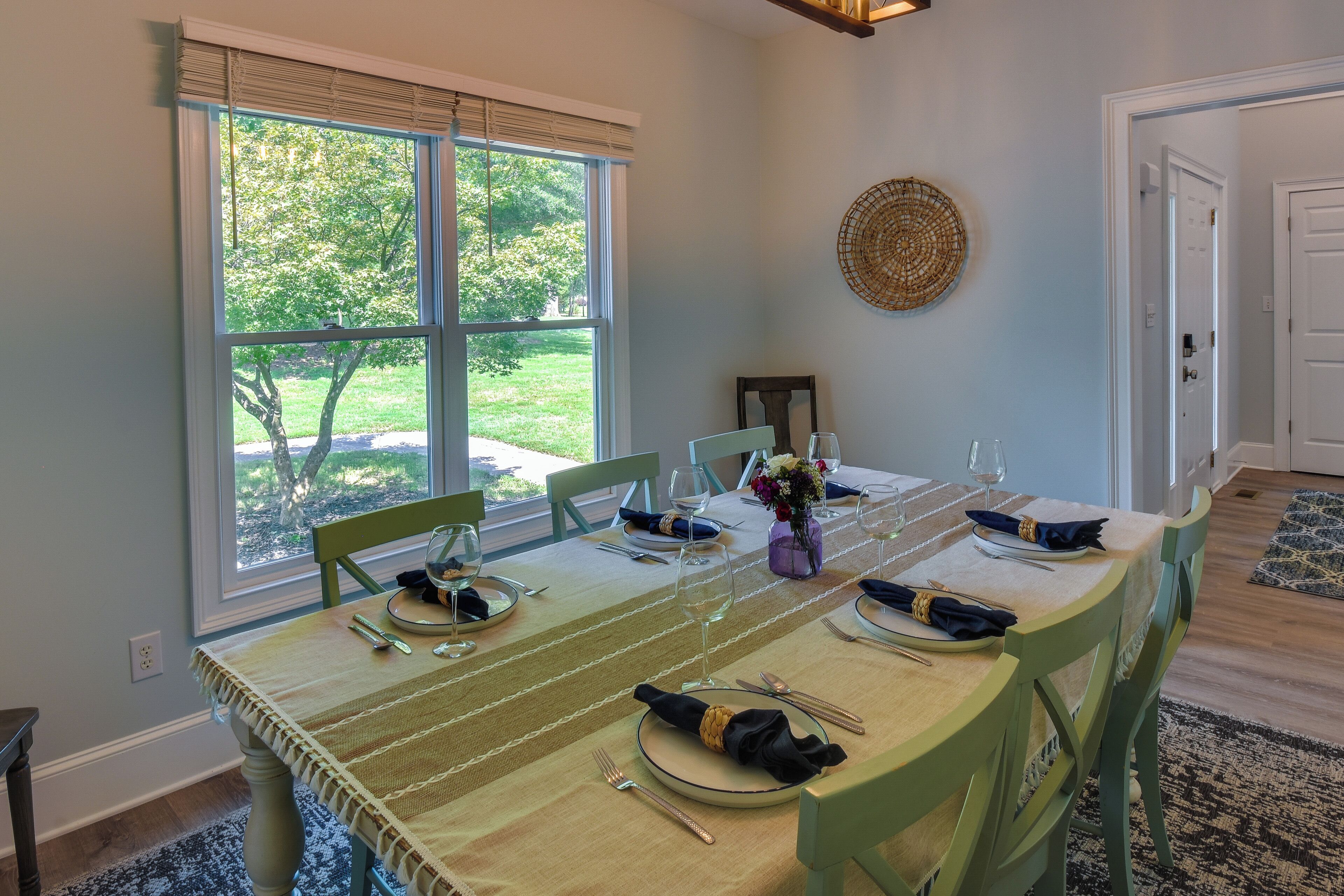 Un comedor con una mesa lista para cuatro personas, una ventana con persianas, un adorno en la pared y una puerta al fondo.