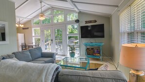 Un salón con un sofá gris, una mesa de centro de cristal, una mesita auxiliar color turquesa, una lámpara y un televisor en la pared.