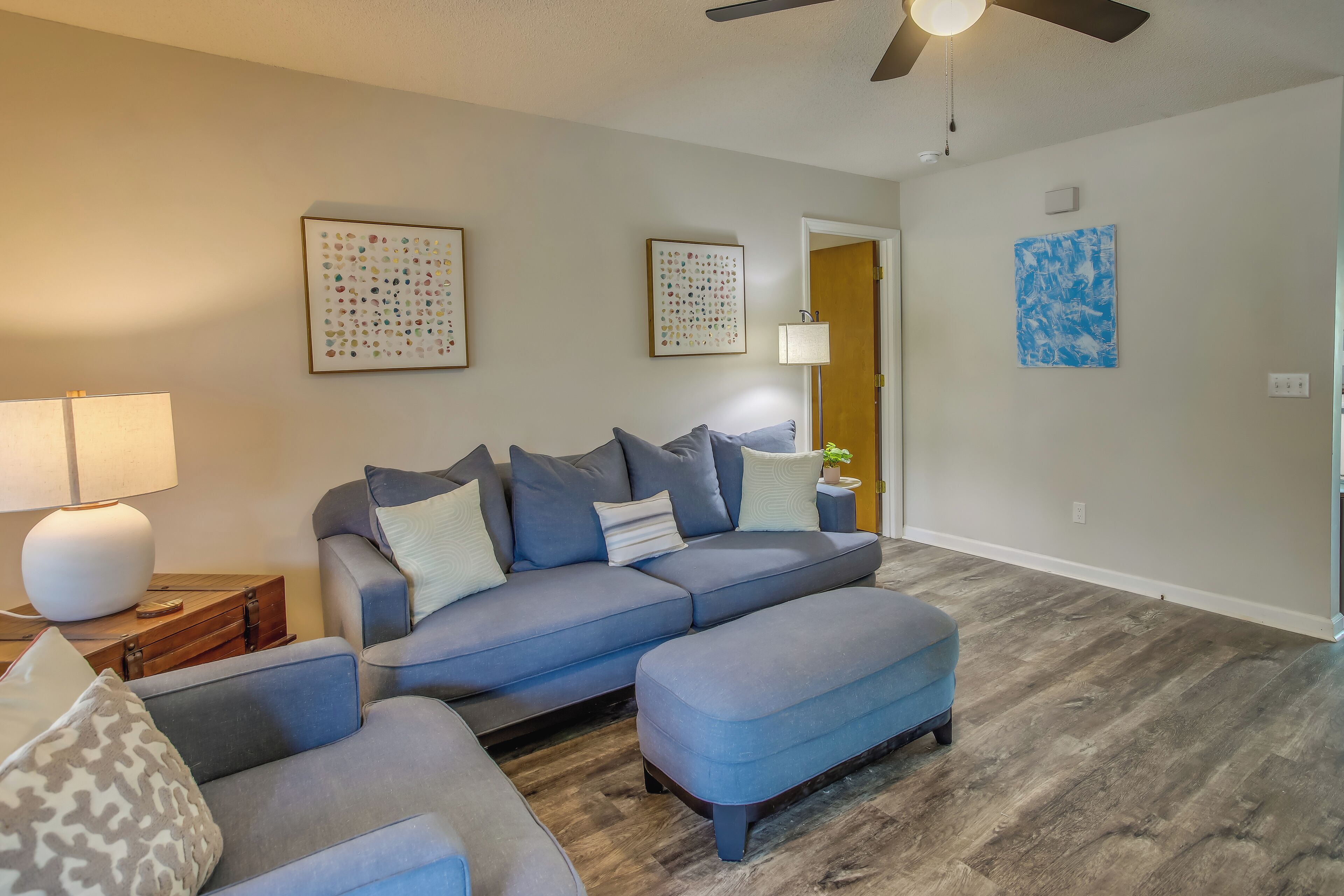 Un salon avec un canapé d’angle gris, un pouf assorti et un plancher en bois. Les murs sont ornés d’œuvres d’art encadrées et un ventilateur de plafond est installé au centre.