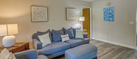 Un salon avec un canapé d’angle gris, un pouf assorti et un plancher en bois. Les murs sont ornés d’œuvres d’art encadrées et un ventilateur de plafond est installé au centre.