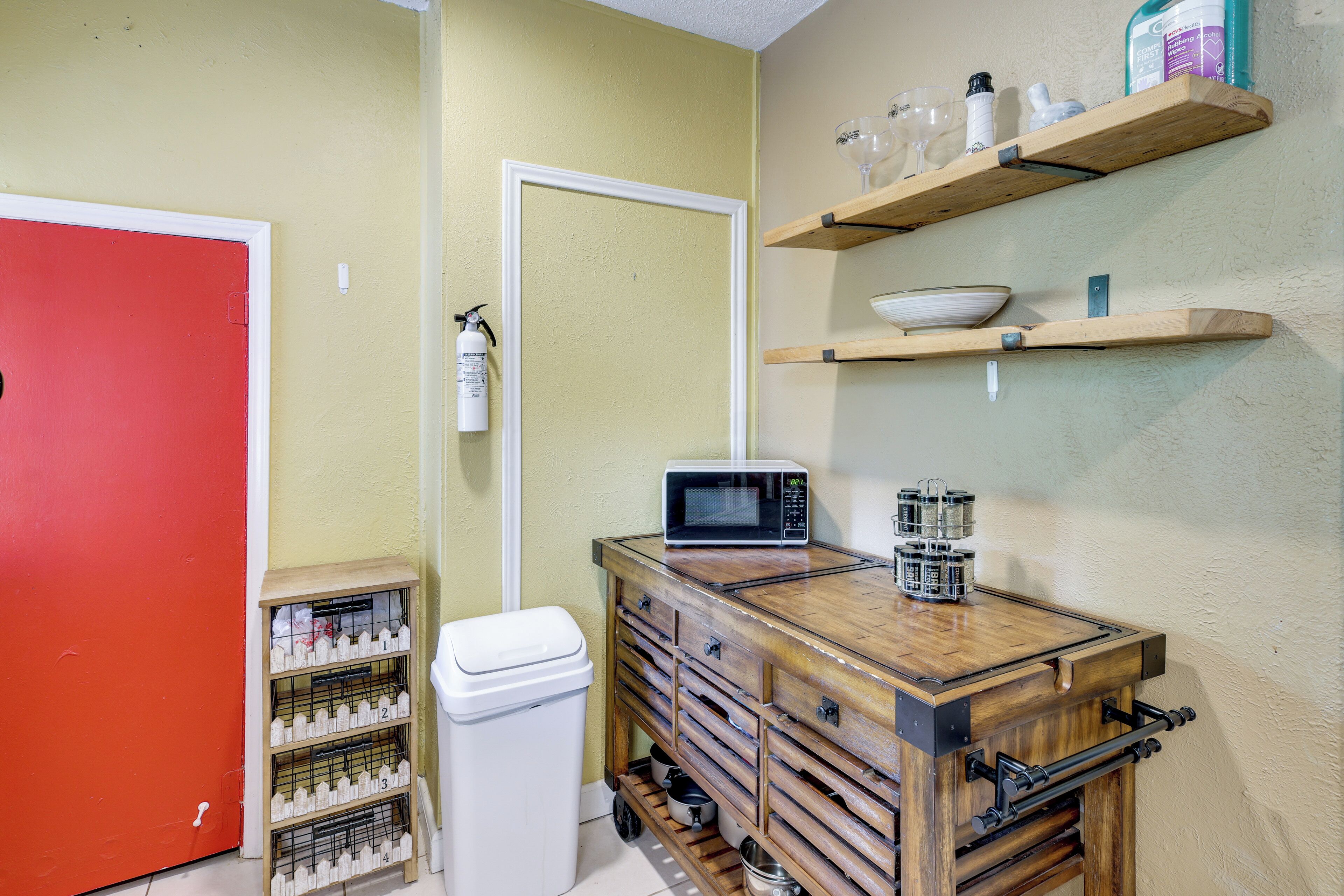 A kitchen with a microwave, shelves with kitchenware, a trash bin, and a red door.