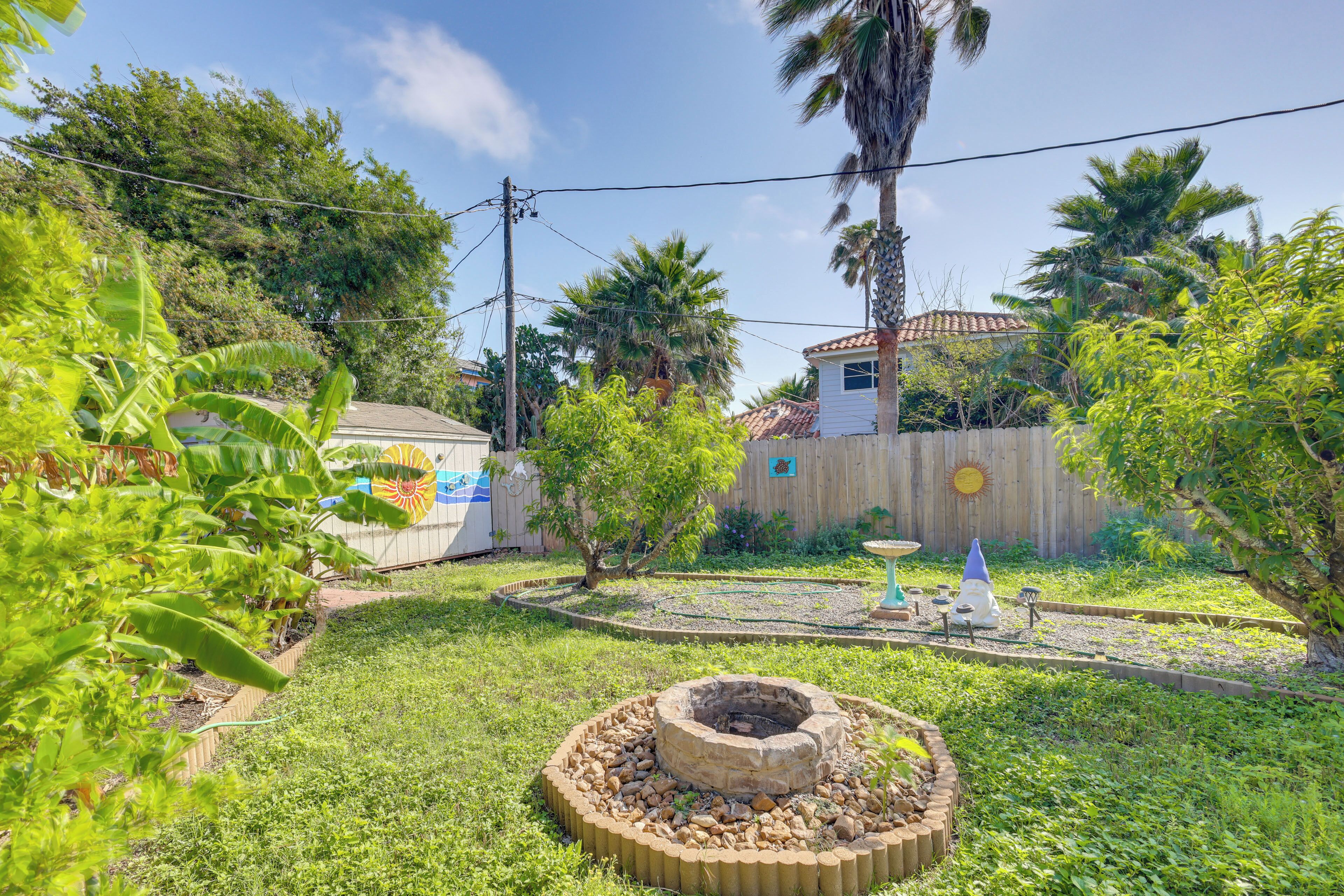 Un jardin avec un foyer circulaire en pierre, une clôture en bois et une maison avec un toit en tuiles.