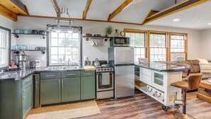 Una cocina con gabinetes verdes, una estufa de acero inoxidable, un microondas y un refrigerador. Hay un taburete de bar y piso de madera.