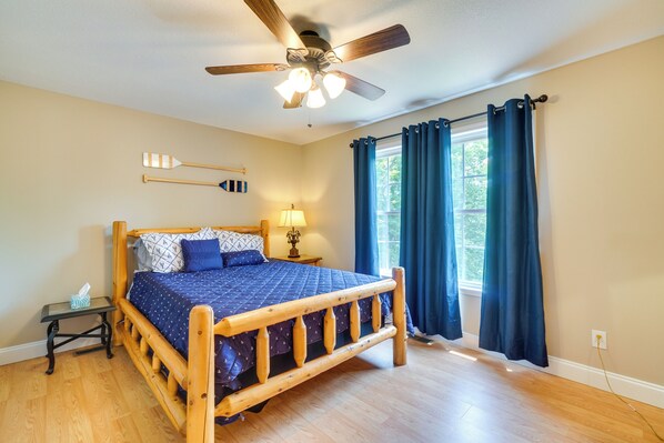 A bedroom with a wooden bed, a ceiling fan, a nightstand, a lamp, and a window with curtains. - Cozy Tennessee Lake House w/ Grill & Fire Pit! (Byrdstown)