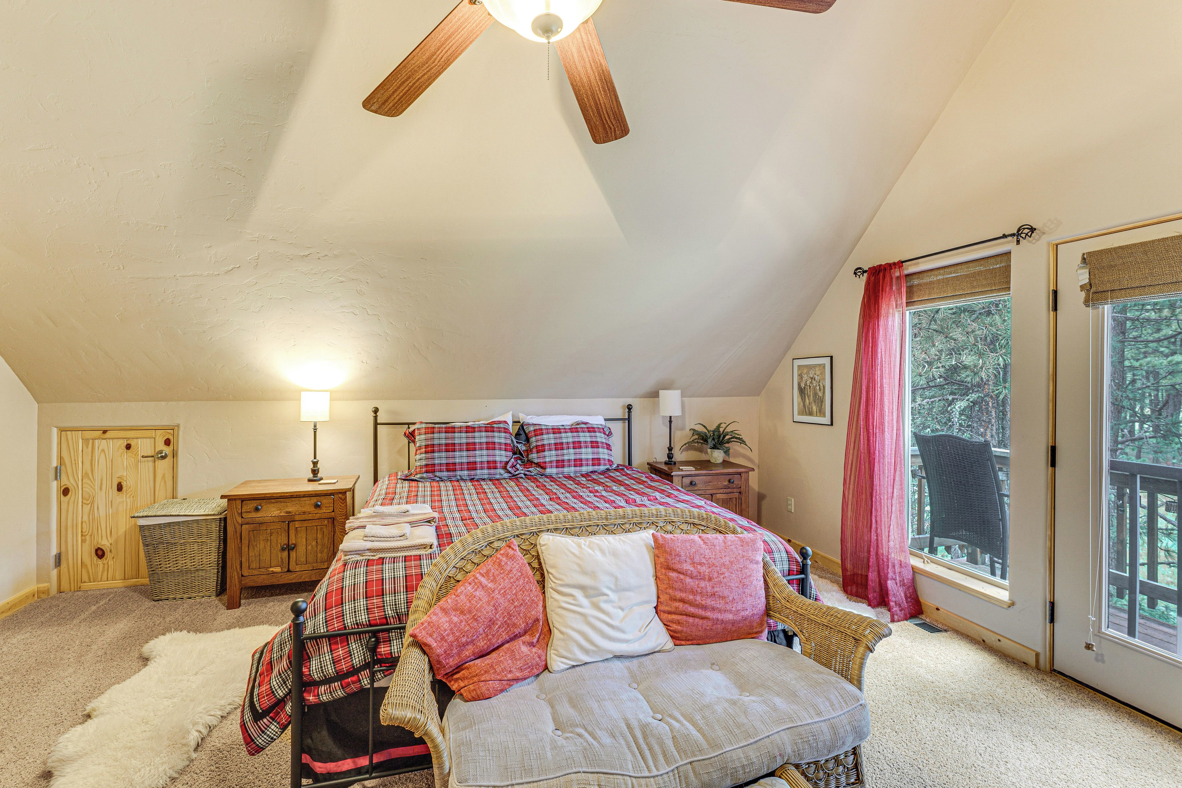 A bedroom with a sloped ceiling, a bed with plaid bedding, a nightstand, a wicker chair, and a view through a window.