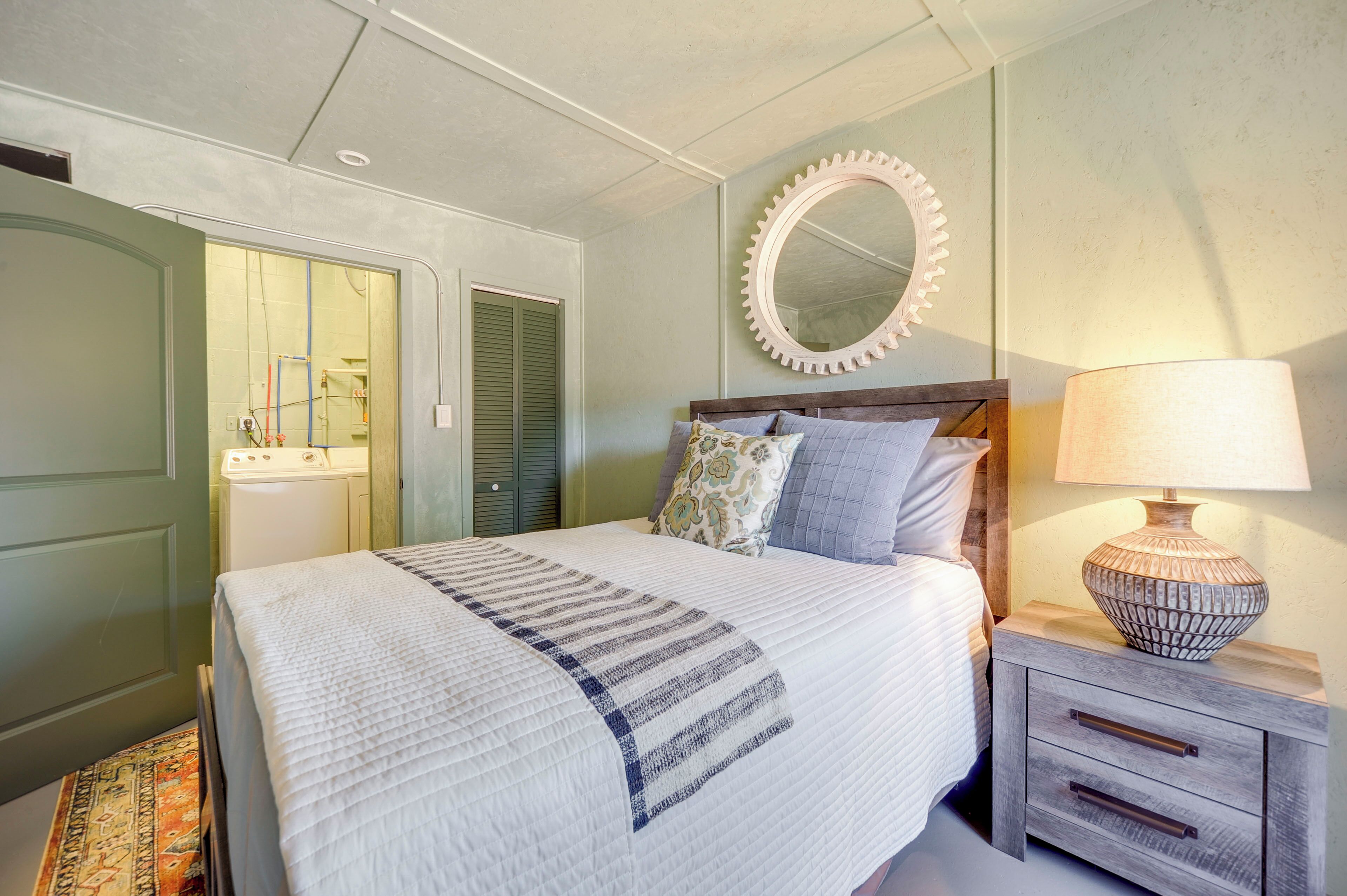 A bedroom with a bed, a nightstand, a lamp, a mirror, and a door leading to a bathroom.