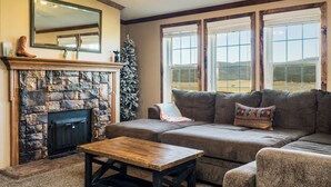 A cozy living room with a stone fireplace, a large window with a view of the outdoors, and a comfortable gray sofa with a decorative pillow. - Angel Fire Home: Sangre de Cristo Mountains View! (Angel Fire)