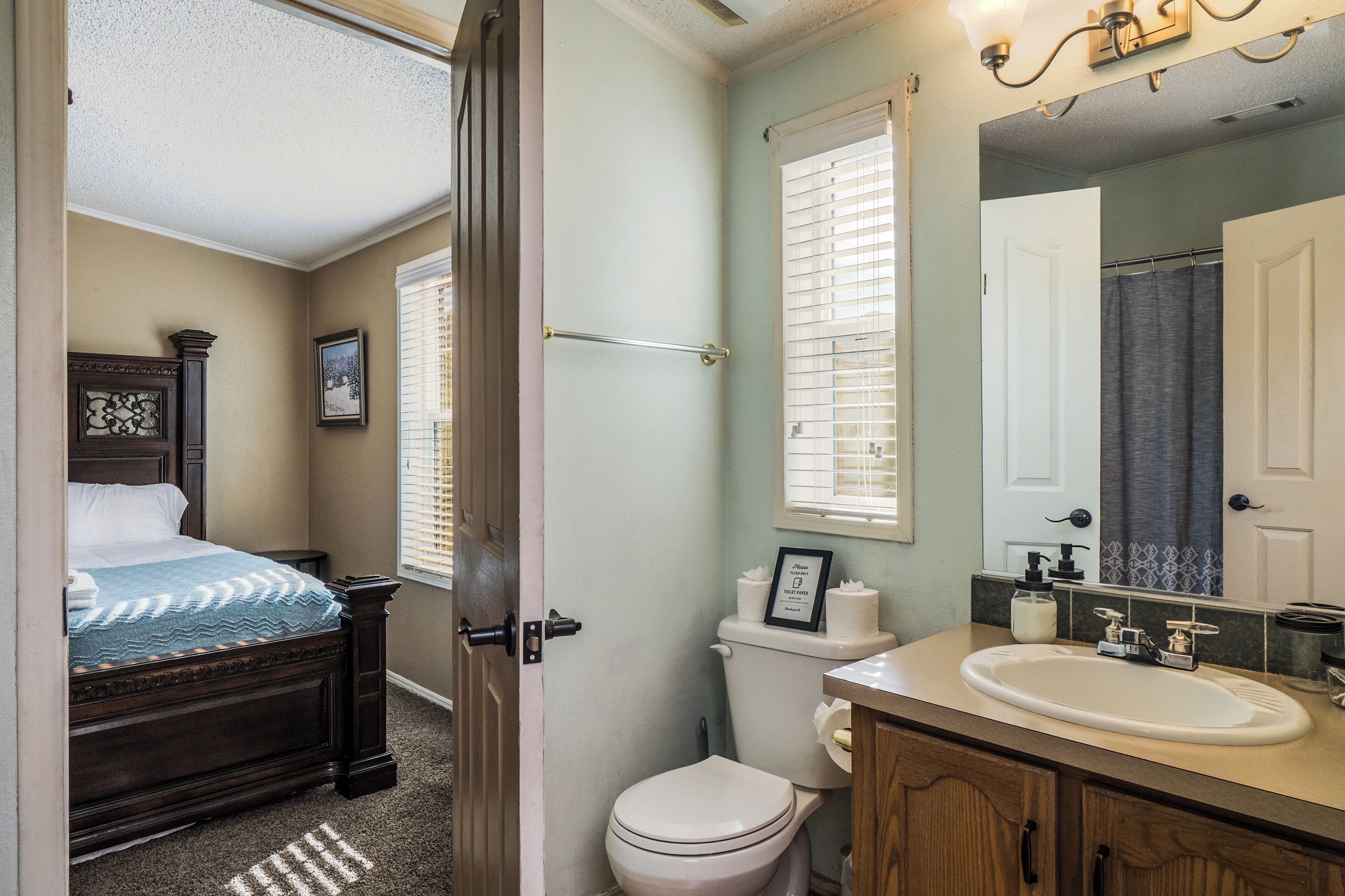 A bathroom with a toilet, sink, and mirror. There is a bed with a headboard and a picture on the wall.