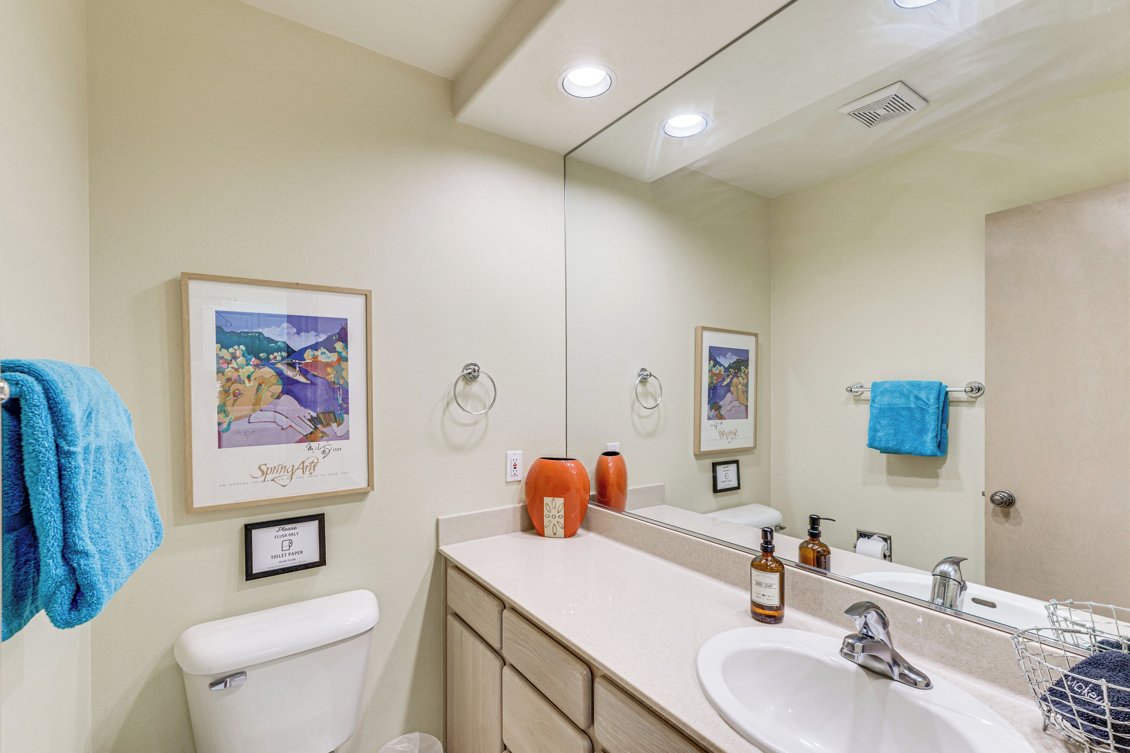 A bathroom with a toilet, sink, and mirror. There are towels hanging on the wall and a framed picture above the sink.