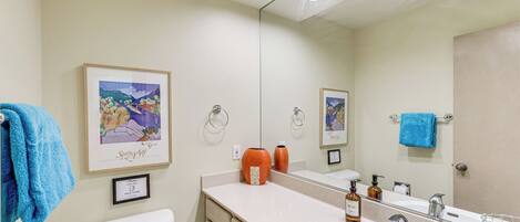 A bathroom with a toilet, sink, and mirror. There are towels hanging on the wall and a framed picture above the sink.