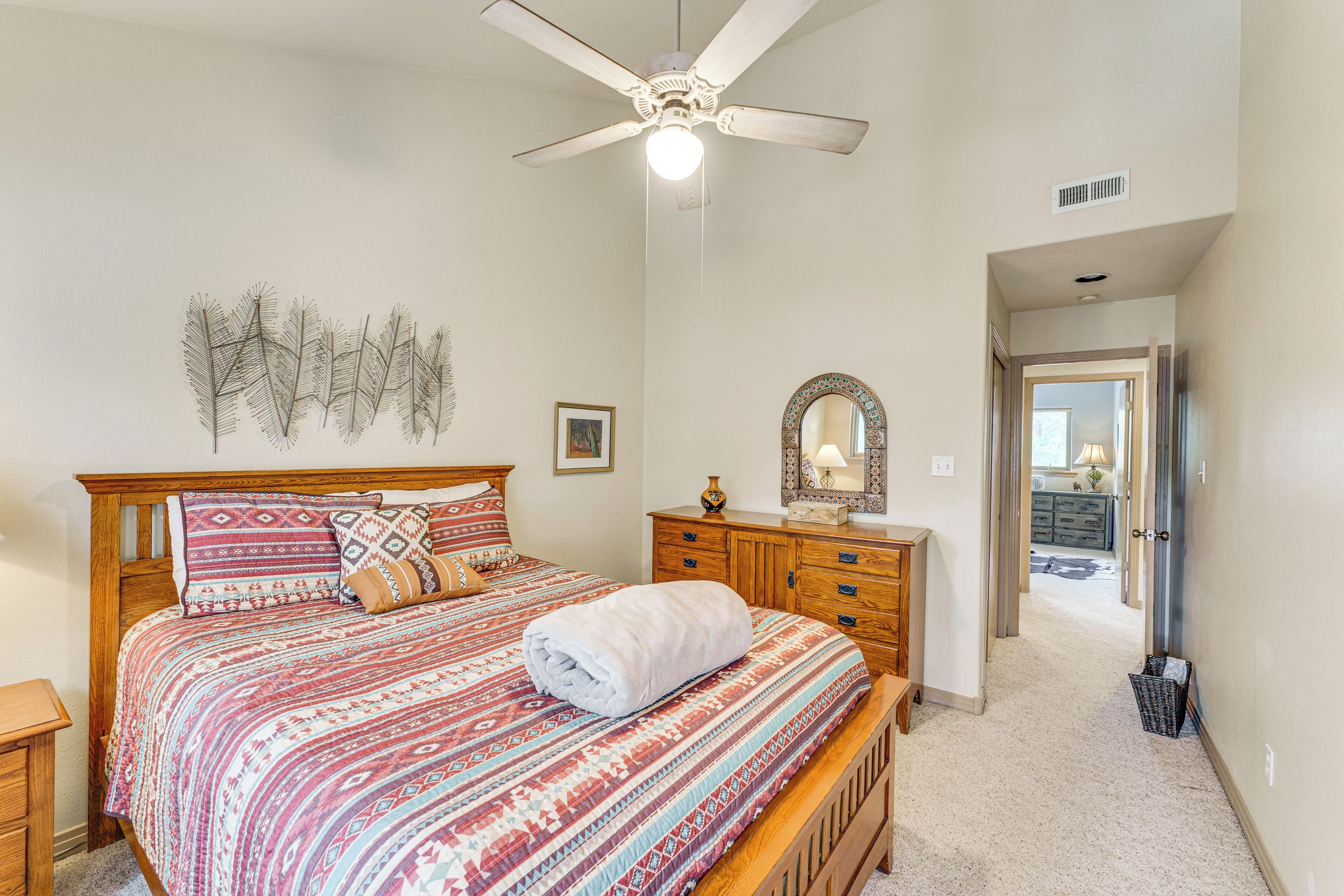 A bedroom with a bed, wooden headboard, a ceiling fan, a dresser, a mirror, and a hallway leading to another room.