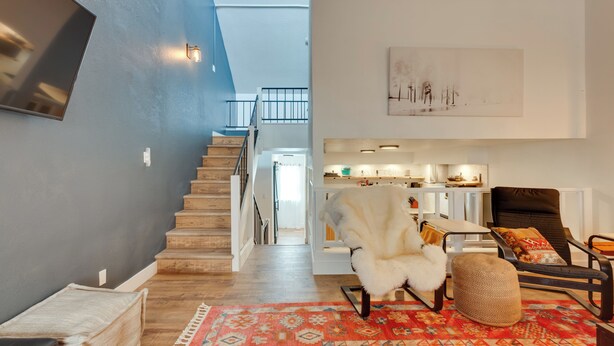 A modern living room with a staircase, a fluffy chair, a patterned rug, and a kitchen area in the background. - Chic Angel Fire Condo: Walk to Ski Resort! (Angel Fire)