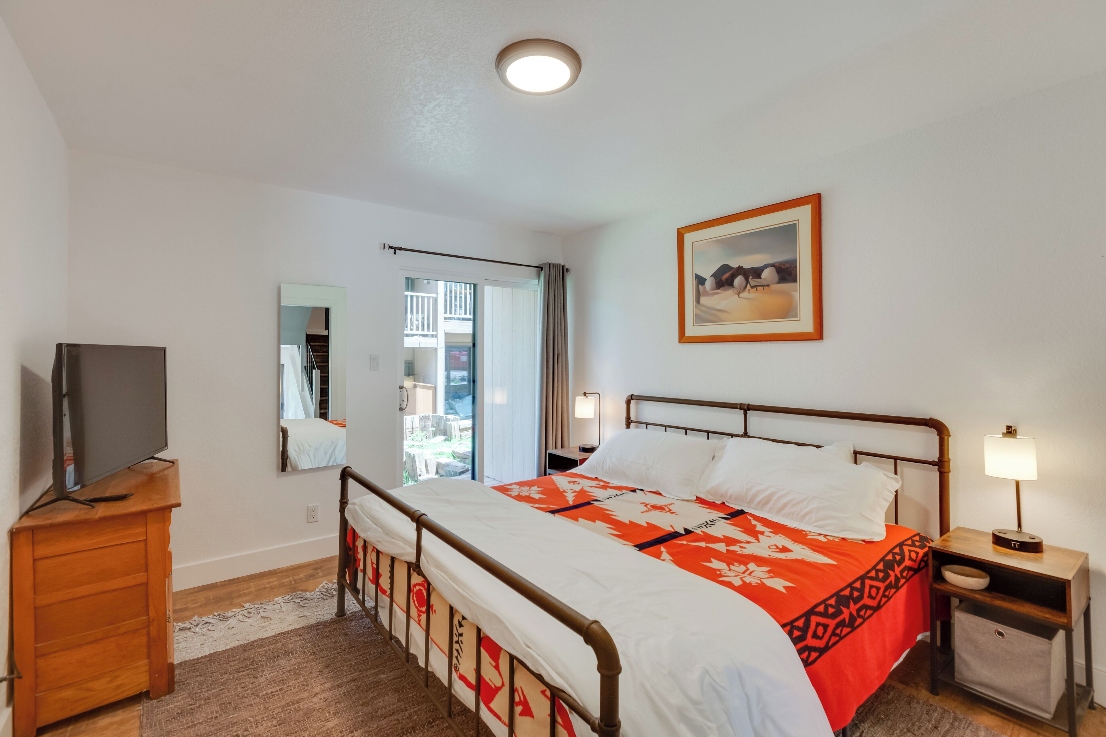 Un dormitorio con cama, televisor sobre un mueble de madera, mesita de noche con lámpara y ventana con cortinas.