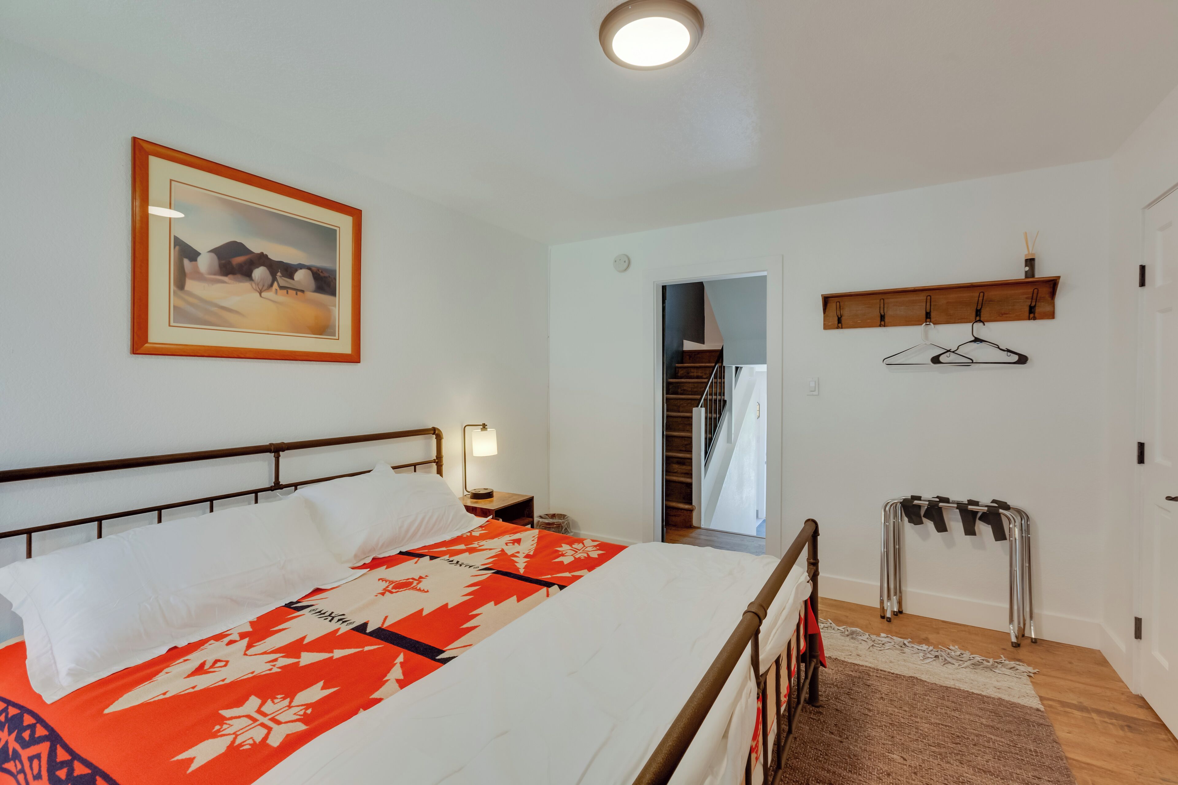 A bedroom with a bed, a nightstand, a wall-mounted shelf, a coat rack, and a door leading to a staircase.