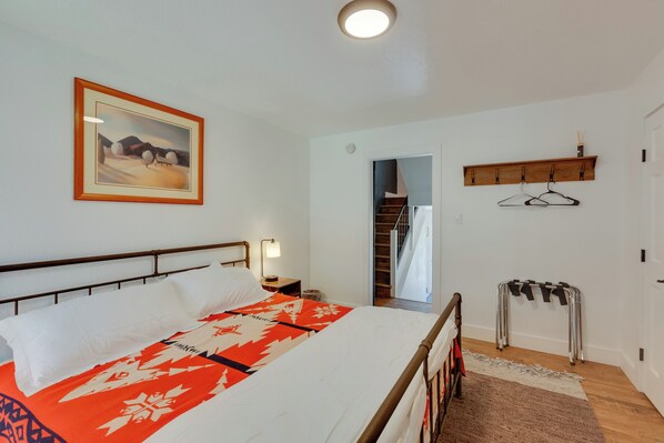 A bedroom with a bed, a nightstand, a wall-mounted shelf, a coat rack, and a door leading to a staircase. - Chic Angel Fire Condo: Walk to Ski Resort! (Angel Fire)